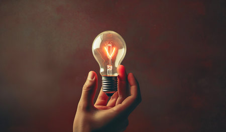 Female hand holding a glowing light bulb on dark red background with copy spaceの素材