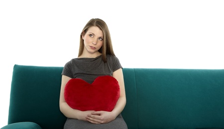 Beautyful young woman smiling and sitting on a sofa with heart shaped pillow in her handの写真素材