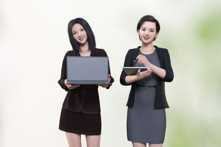 A female employee holds a laptop computer in her hand, with the screen facing us, and a female...
