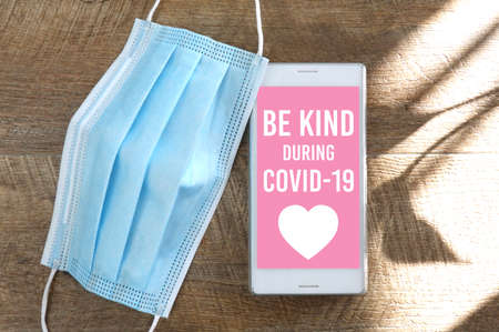 Coronavirus or Covid-19 be kind campaign on screen, Smartphone and face mask on wooden table