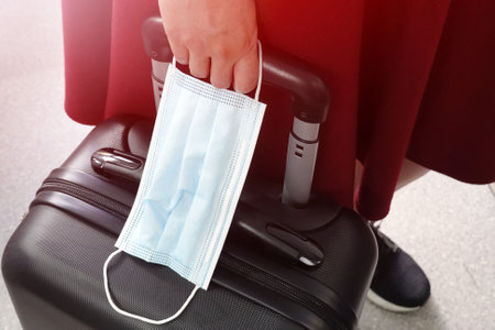 Travelling during covid-19 pandemic. Woman holding protective mask and black suitcaseの写真素材