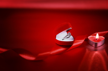 Wedding solitaire ring in heart shaped red box with candle and red ribbon on red background....