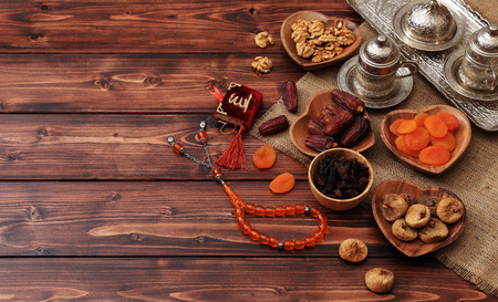 Ramadan concept. Delicious dates, dried figs, dried apricots, walnuts, almonds, raisins on bamboo and silver plates, rosary beads with Islamic holy book Quran on a pine wooden table background.の写真素材