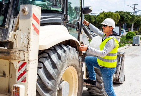 Portrait of a male worker in a white helmet and reflective vest operating a tractor at a...
