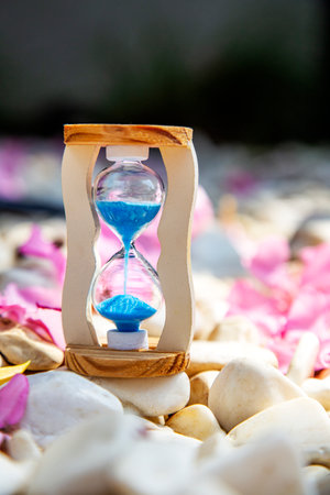 Hourglass with blue sand on white stones and pink oleander petals nature background. Life time,...