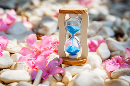 Hourglass with blue sand on white stones and pink oleander petals nature background. Life time, meaning of life and beauty passing conceptの写真素材
