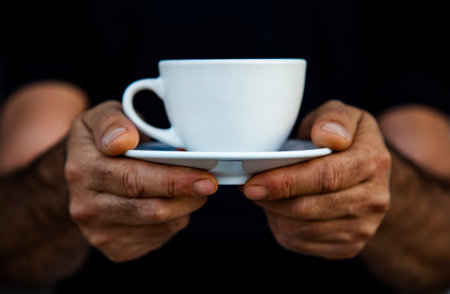 Close-up of man hands holding white cup of coffee on black background. the mug with hot drink on a saucerの写真素材