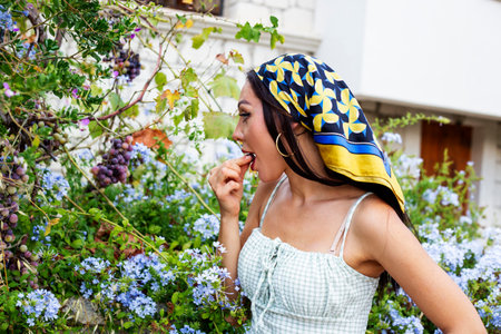 Portrait of a beautiful young brunette woman in a headscarf in the garden eating grapes