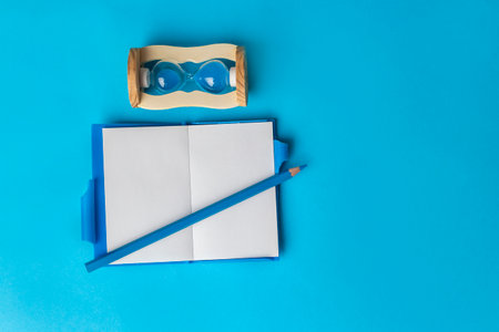 Notebook, pencil and hourglass on a blue background. Top view copy space