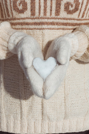 A girl in a beige sweater holds in her hands, dressed in white mittens, a heart made of snow close-up. Postcard for Valentine's Dayの写真素材