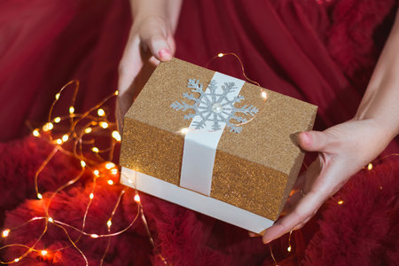 A gift in women's hands on the background of a red dress with lights from a garlandの写真素材