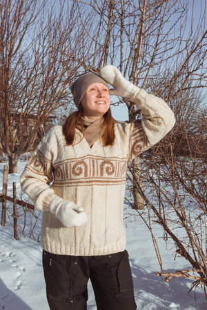 A happy woman in a beige patterned sweatshirt and a hat looks up against the background of a winter landscapeの写真素材