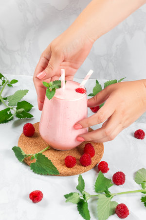 Women's hands put a milkshake with raspberries in a glass glass on the tableの写真素材