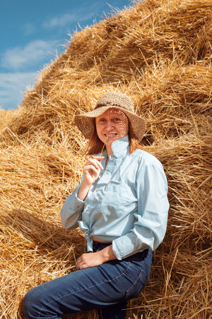 A woman in a shirt, jeans and a straw hat leans on a haystack and chews a spikeletの写真素材