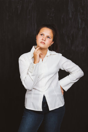 A schoolgirl in a white shirt on a black background stands with a thoughtful lookの写真素材