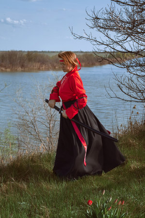 A woman with short red hair in black and red hanfu and a katana on the background of a river in springの写真素材