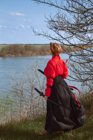 A woman with short red hair in a black and red hanfu and two katanas looks into the distance against the background of a spring riverの写真素材