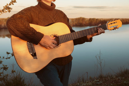 A woman in a warm sweater and pants plays a classical guitar against the background of a river at...