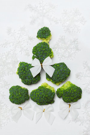 Green broccoli lie in the shape of a Christmas tree with white bows and snowflakes on a white backgroundの写真素材