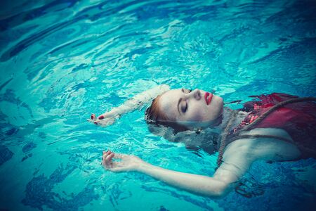 Young sexy woman floating on swimming pool in red dress. Beautiful rich lady who was strangled and thrown off into the waterの写真素材