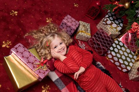 Happy small girl in dress with present have a christmas. Happy little girl holding gift box by christmas tree. Red and goldenの写真素材