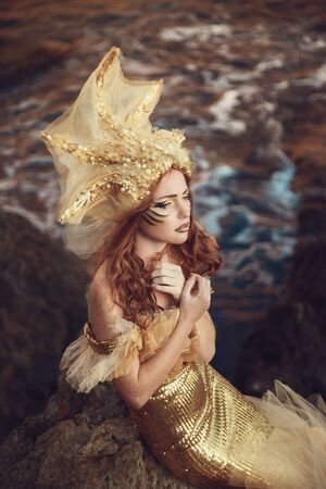 Beautiful fashionable mermaid with red hair and golden scales sitting on a rock by the seaの写真素材