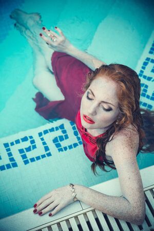 Young sexy woman floating on swimming pool in red dress. Beautiful rich lady who was strangled and thrown off into the waterの写真素材