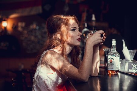 Sad sexy woman sitting on the bar counter at night with glass of whiskeyの写真素材