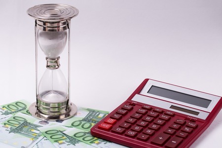 The hourglass is on Euro banknotes and the calculator on a light background. The concept of time is...