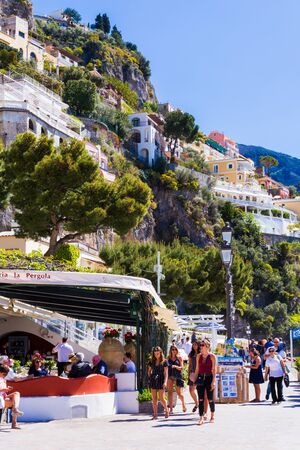 Positano, Amalfi Coast, Italy - 9 April 2017: Beautiful young girls walking along the embankment of the city and other tourists having a rest in the restaurant on a sunny afternoon.のeditorial素材