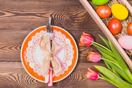Top view of orange plate and cutlery,easter colorful eggs on a bed of straw in a long wooden box on a wooden table and red tulips.の写真素材