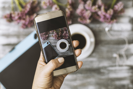 Blogger photographer workshop concept, hand holding a phone and taking photos of stylish flowers, a cup of coffee and a tablet computer on a gray wooden rustic backgroundの写真素材