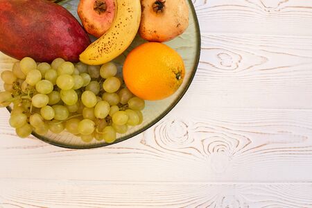 Juicy fruit grapes, banana, mango, pomegranate, oranges on a plate on a wooden white table. Copy space, flat lay.の写真素材