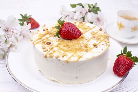 Cake with cream and nuts on a white plate, juicy strawberries and a cup of coffee and flowers on a...