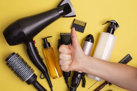 Hairdressing tools in black: hair dryer, combs, sprays, clipper and female hand with thumbs up on a yellow background, flat lay.の写真素材