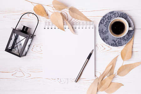 Top view of an empty scrapbook and a metal pen, a cup of coffee, beige leaves, a black iron...