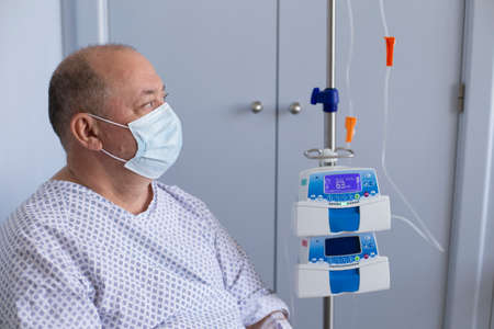 Overweight man wearing a medical mask sits in a chair with a dropper, equipment in the foreground, patient in hospital.の写真素材