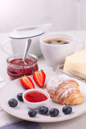 Breakfast with fresh croissant and jam, butter, ripe strawberries and blueberries and a cup of coffee on a white table, vertical photo.の写真素材