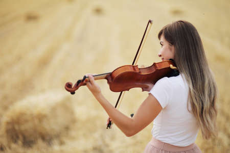Beautiful romantic girl with loose hair playing the violin in the field after the harvest. Square sheaves of hay in the field. Violin trainingの写真素材