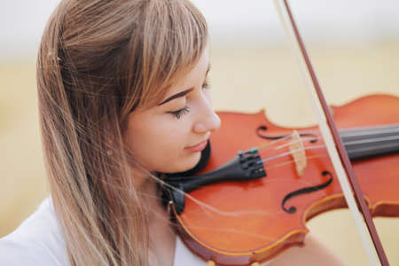 Beautiful romantic girl with loose hair playing the violin in the field after the harvest. Square sheaves of hay in the field. Violin trainingの写真素材