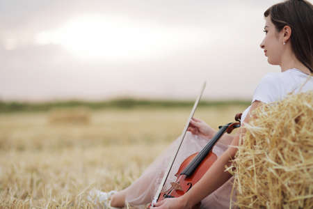 Romantic young woman with flowing hair, holding a violin in her hand in a field after harvest. Square sheaves of hay in the field. Violin trainingの写真素材