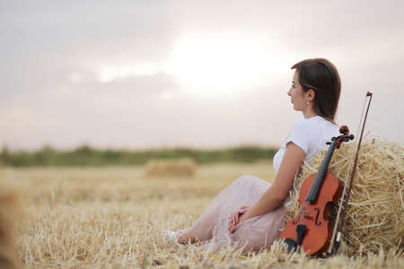 Romantic young woman with flowing hair, holding a violin in her hand in a field after harvest. Square sheaves of hay in the field. Violin trainingの写真素材