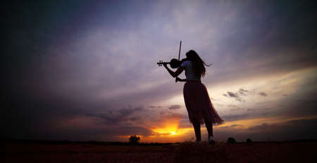 Romantic young woman with loose hair playing the violin in a field at sunset after harvest. Square sheaves of hay in the field. Violin trainingの写真素材