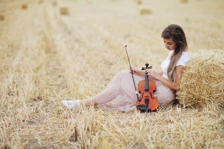 Romantic young woman with flowing hair, holding a violin in her hand in a field after harvest. Square sheaves of hay in the field. Violin trainingの写真素材
