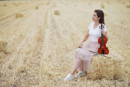 Romantic young woman with flowing hair, holding a violin in her hand in a field after harvest. Square sheaves of hay in the field. Violin trainingの写真素材