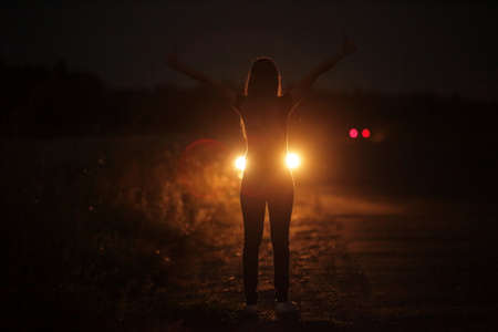 Silhouette of young slender woman in the backlight of car headlights on the auto road at night. The girl is hitchhiking.の写真素材