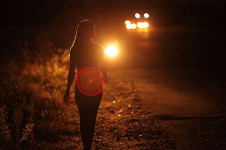Silhouette of young slender woman in the backlight of car headlights on the auto road at night. The girl is hitchhiking.の写真素材