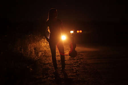 Silhouette of a slender young woman with violin in car headlights illumination. A young slender girl in the backlight of car headlights on the auto road at night. The girl is hitchhiking.の写真素材
