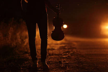 Silhouette of a slender girl's legs and a violin in the backlight of headlights. A young slender woman in the backlight of car headlights on the auto road at night. The girl is hitchhiking.の写真素材