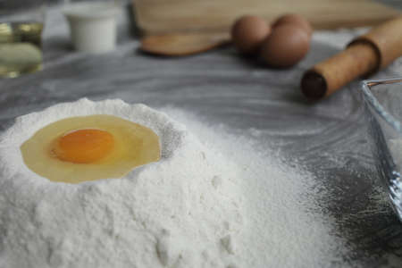 Broken chicken egg in a pile of flour, olive oil, milk, kitchen tool on gray table background. Products for baking bakery products. Cutting board, rolling pin, flour sieve, wooden spoon. bread or cakeの写真素材